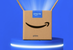 The image shows a cardboard box with the Amazon logo and the words "prime day" on a blue label. The box is open and placed on a blue platform with a glowing edge, set against a blue background with light beams emanating from the box.
